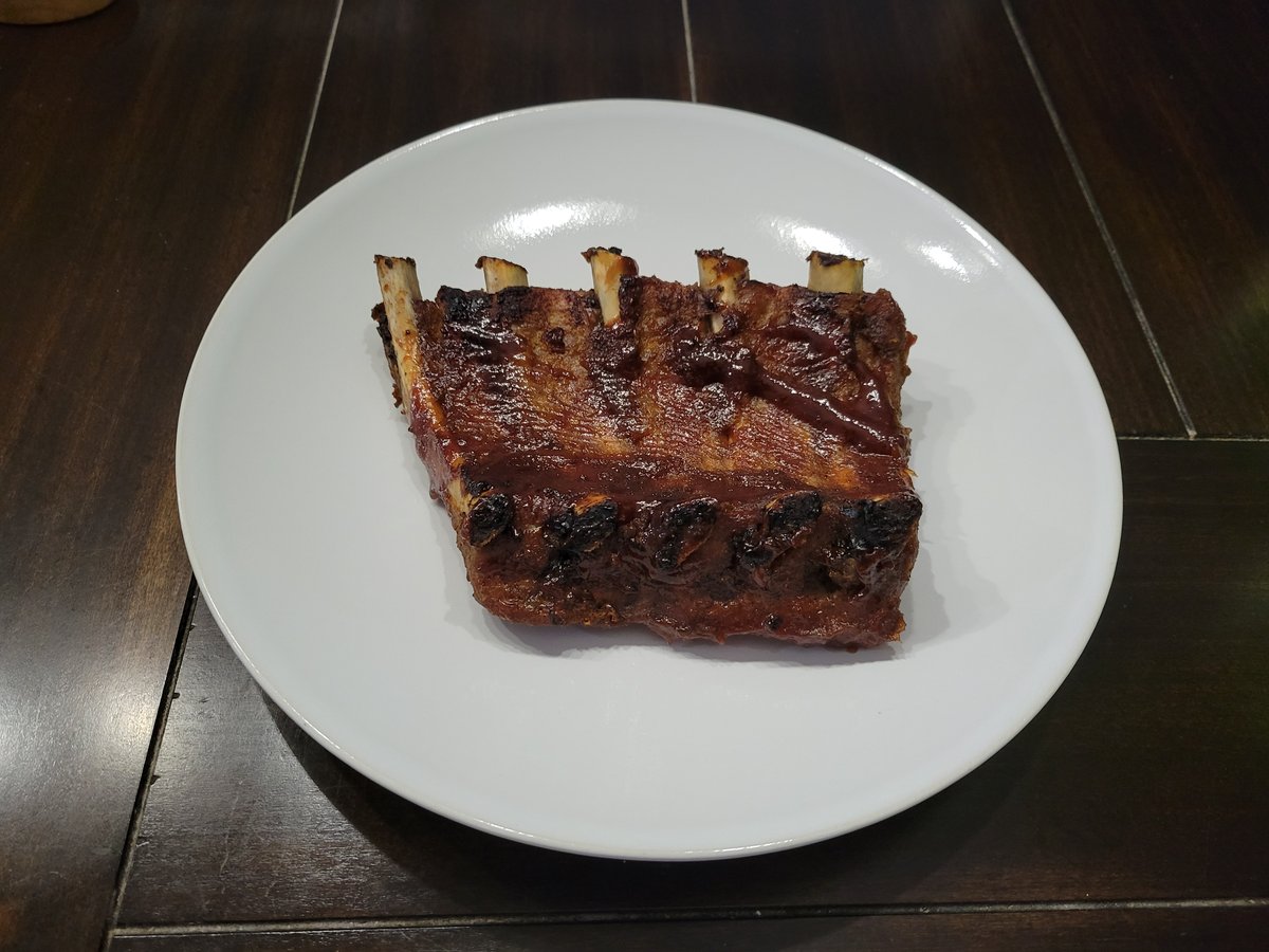A mahogany-glazed half-rack of babyback ribs on a white plate, glossy BBQ sauce, cleanly cut bones exposed, a little char on the ends