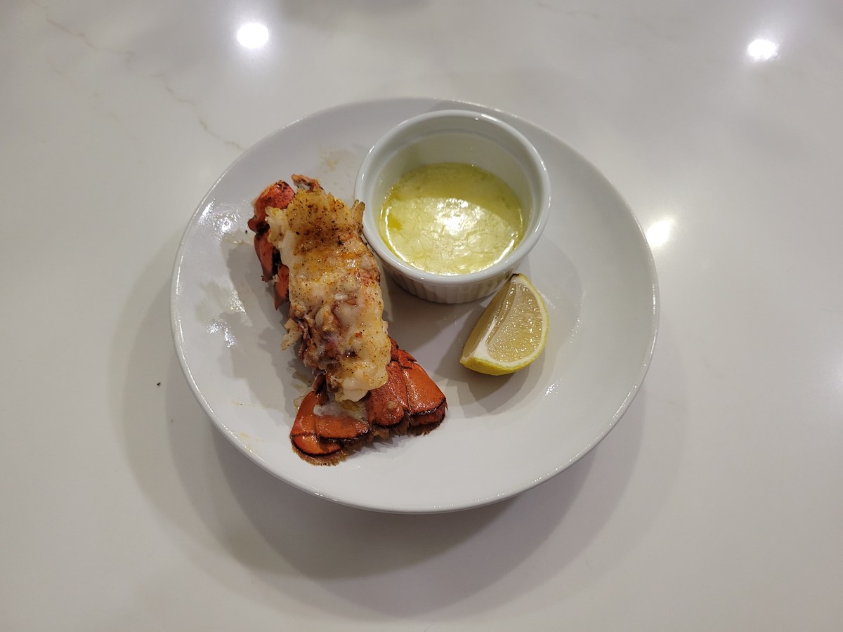 A single baked lobster tail butterflied on top of its red shell, dusted with paprika, served with a ramekin of melted butter and a lemon wedge on a white plate.