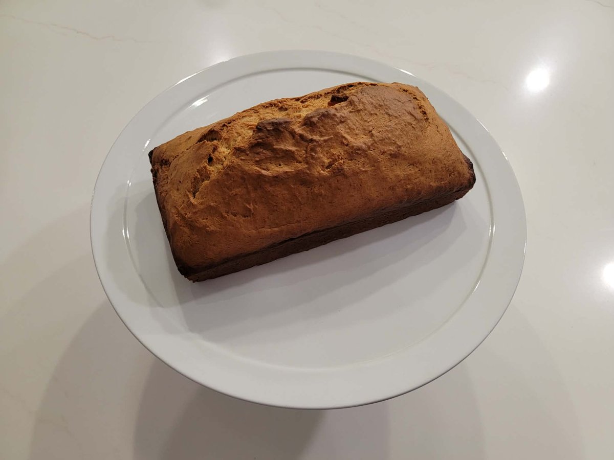 A golden-brown loaf of banana bread on a white plate, with a clean crack down the center of the top crust.
