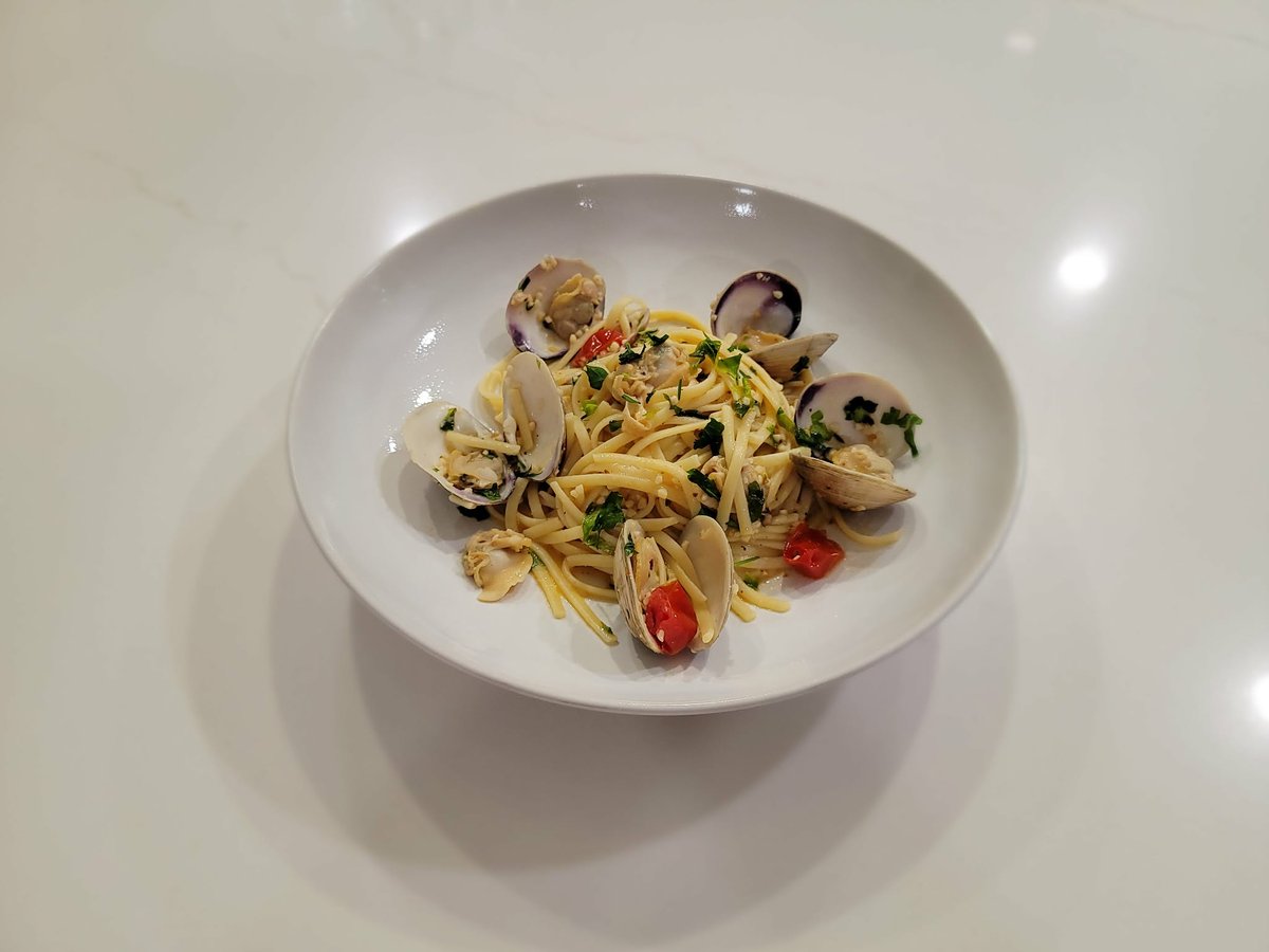 A shallow white bowl of linguine alla vongole on a marble surface, tangled with open clam shells showing the meat, a few closed shells, halved grape tomatoes peeking between the noodles, and a scatter of chopped parsley across the top.