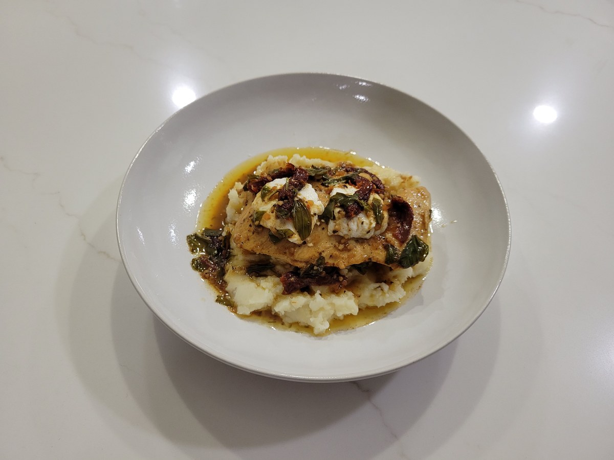 Plated Chicken Bryan: a golden-brown chicken breast set on a mound of mashed potatoes, topped with soft goat cheese, sliced sun-dried tomatoes, and torn fresh basil, with glossy lemon-butter sauce pooling around the base in a wide shallow white bowl.
