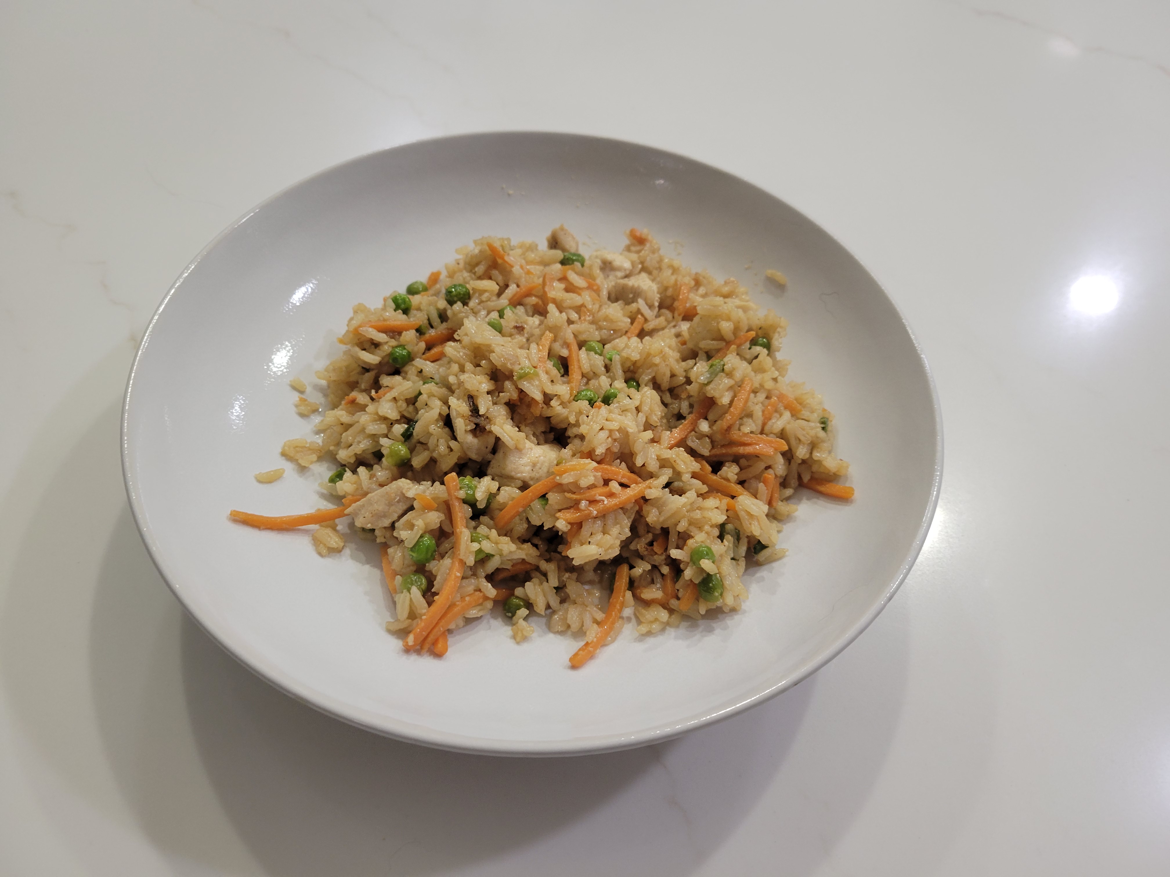A wide bowl of chicken fried rice, golden-brown grains, diced chicken, matchstick carrots, and green peas throughout. No egg on top.