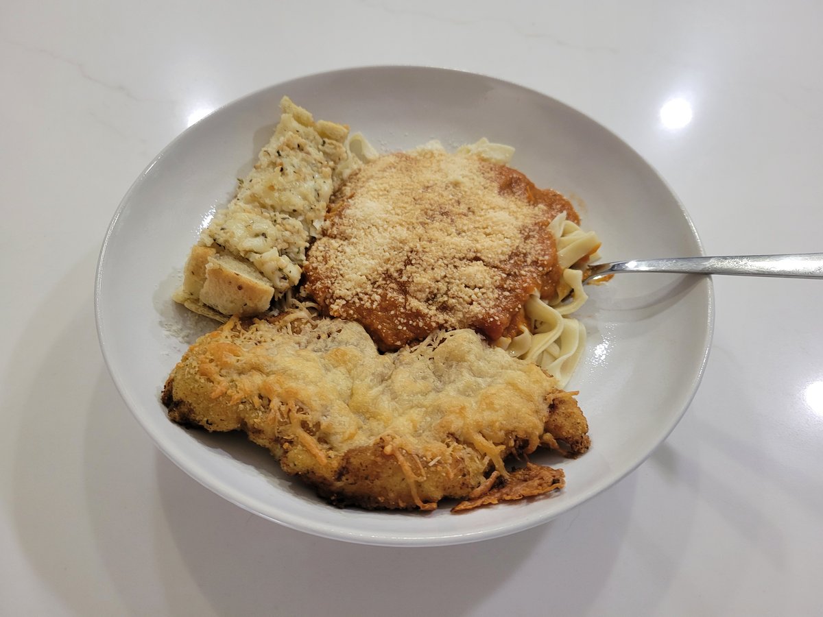 A white bowl of chicken parmesan: a breaded chicken cutlet topped with melted parmesan, on a bed of spaghetti in red sauce dusted with parmesan, with a slice of garlic bread alongside and a fork stuck in.