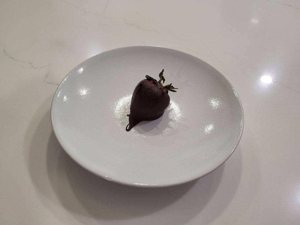 A single chocolate-covered strawberry on a large white plate, with a glossy set semi-sweet chocolate coating and the green leaves left on