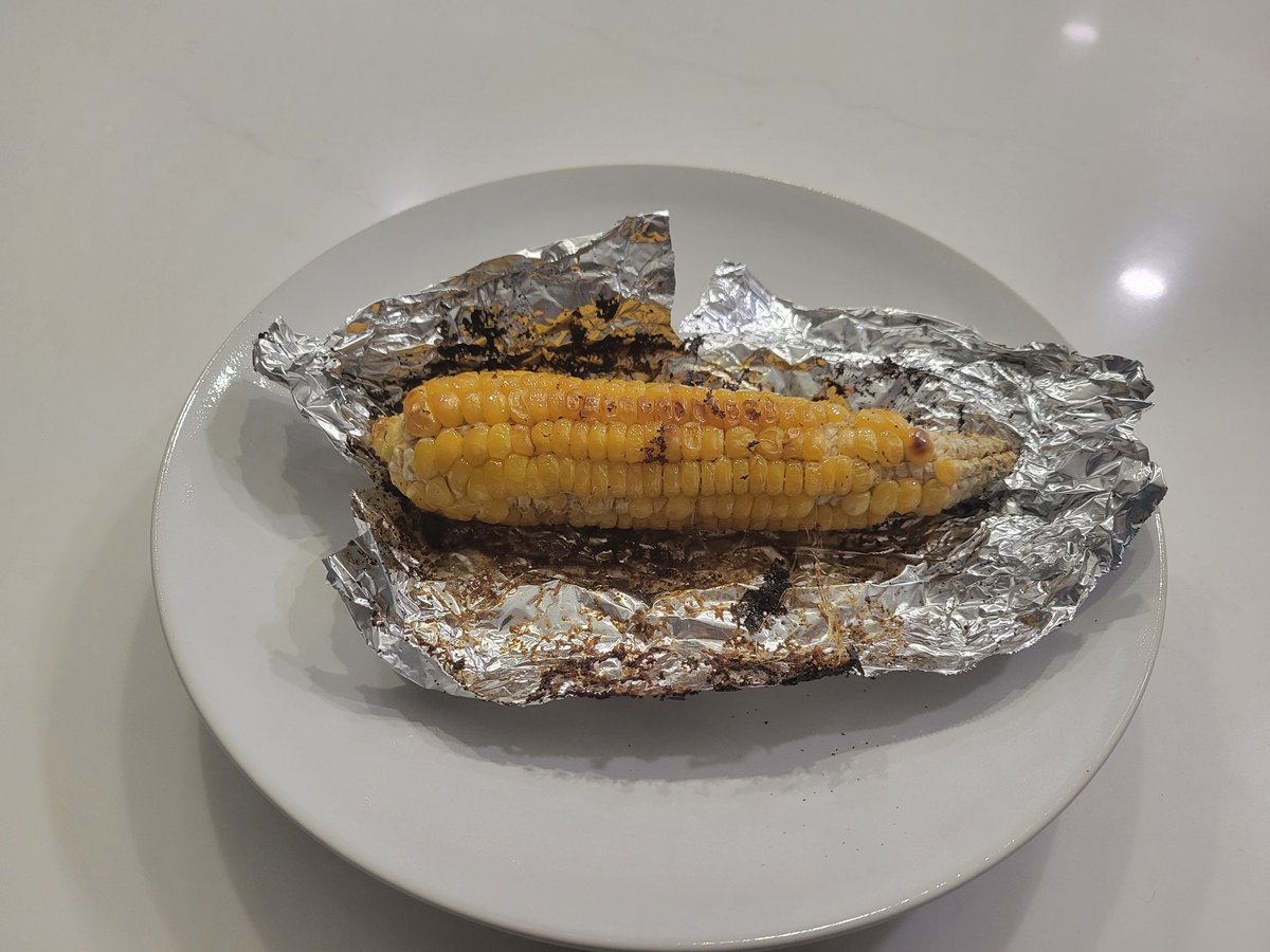 A grilled ear of corn on a plate, foil peeled back, kernels lightly charred and butter pooled in the open foil