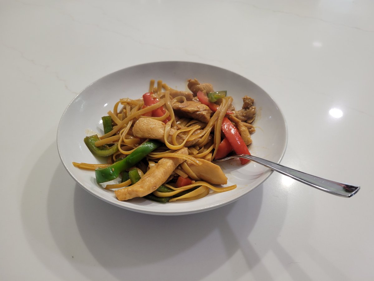 A bowl of faux mein: chicken stir fry with red and green bell peppers over fettuccine noodles in a glossy brown sauce.