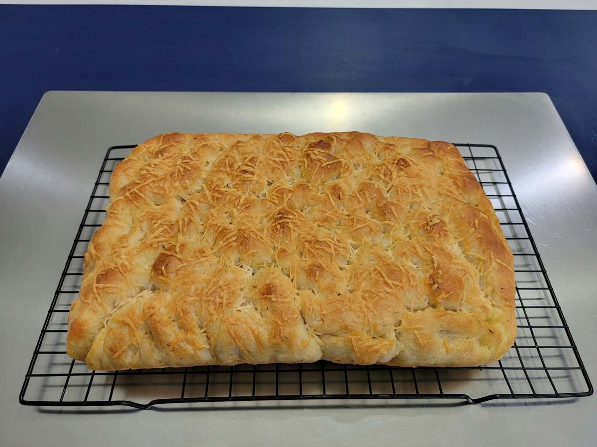 Golden-brown focaccia on a black cooling rack over a stainless steel sheet pan, with visible fingertip dimples and browned parmesan across the top