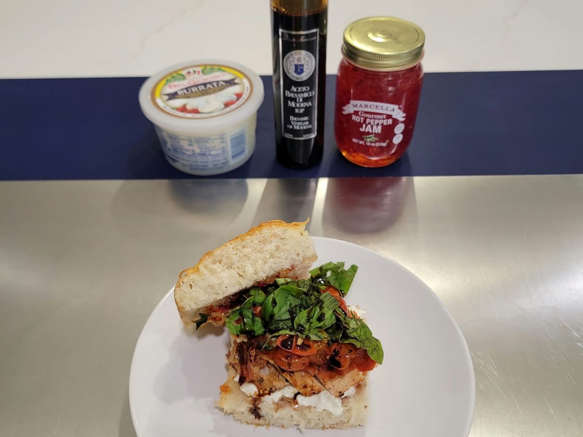 A focaccia sandwich on a white plate with burrata, crispy prosciutto, roasted tomatoes, basil and balsamic glaze, with BelGioioso burrata, Aceto Balsamico di Modena IGP and Marcella Gourmet hot pepper jam on the counter behind it