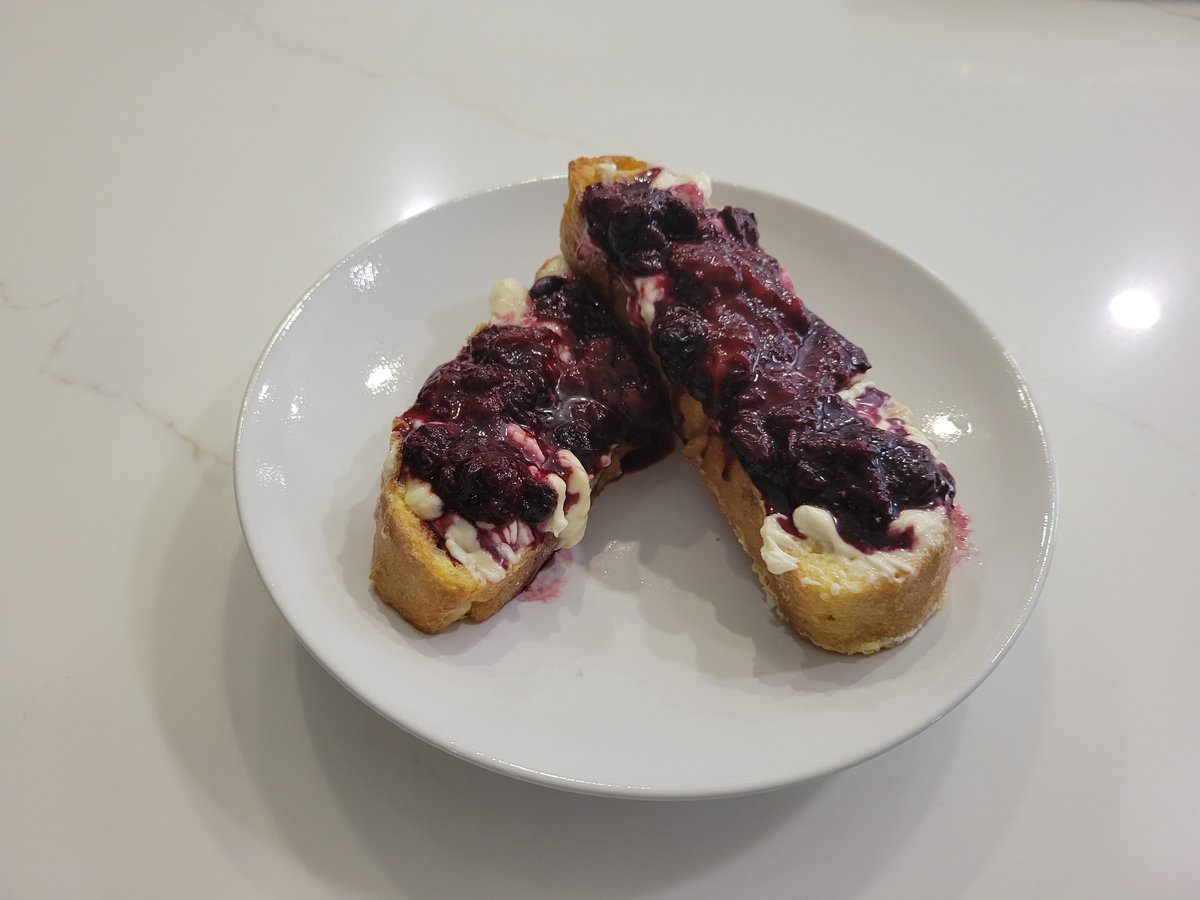 Two golden French toast planks topped with cream cheese and dark purple mixed-berry compote on a white plate