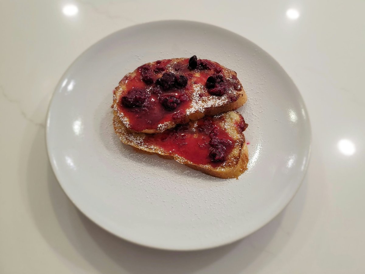 Two slices of golden French toast on a white plate topped with deep purple-red mixed berry compote and a dusting of powdered sugar
