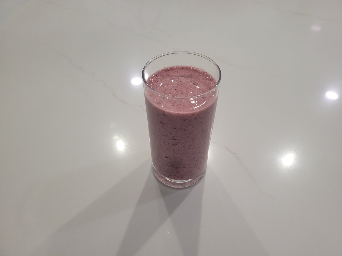 A tall clear glass of thick, dusty pink-purple fruit smoothie with visible flecks of blueberry skin