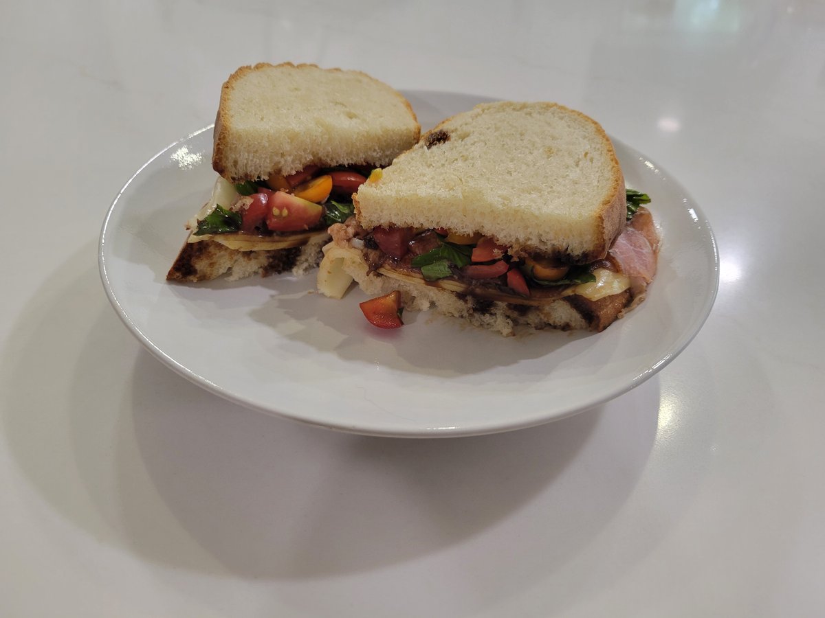 Two halves of an Italian sandwich on a white plate: soft rustic bread with a little char on the bottom crust, fresh mozzarella, prosciutto, chopped red and yellow heirloom tomatoes, basil, and dark balsamic glaze drizzled through the filling