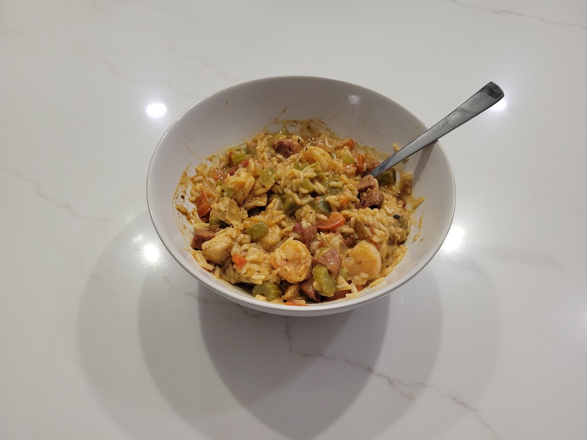 A white bowl of jambalaya with chicken, sliced andouille sausage, pink shrimp, diced peppers, celery, and visibly separate grains of rice, with a spoon sticking out.