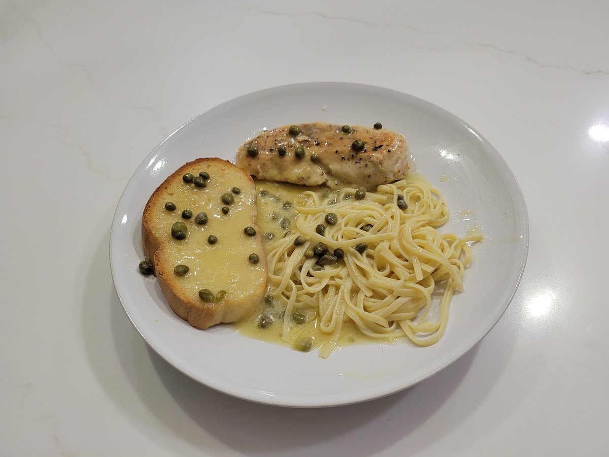 A plate of chicken piccata: a pan-seared chicken breast in glossy lemon-butter sauce dotted with bright green capers, alongside linguine and a slice of bread, with the sauce pooling around the plate.