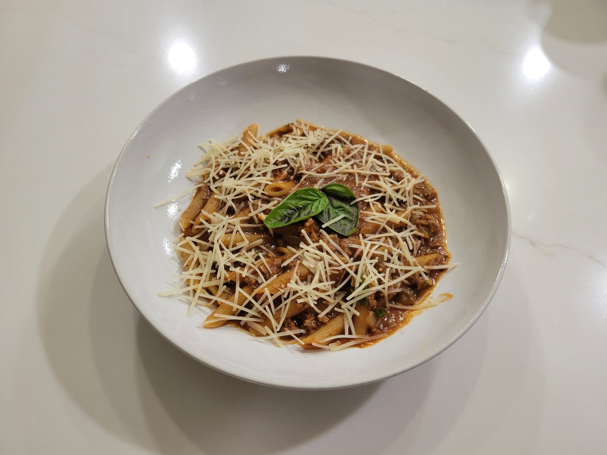 A wide bowl of penne in a glossy brick-red beef and tomato sauce, mounded with shredded parmesan and two fresh basil leaves on top