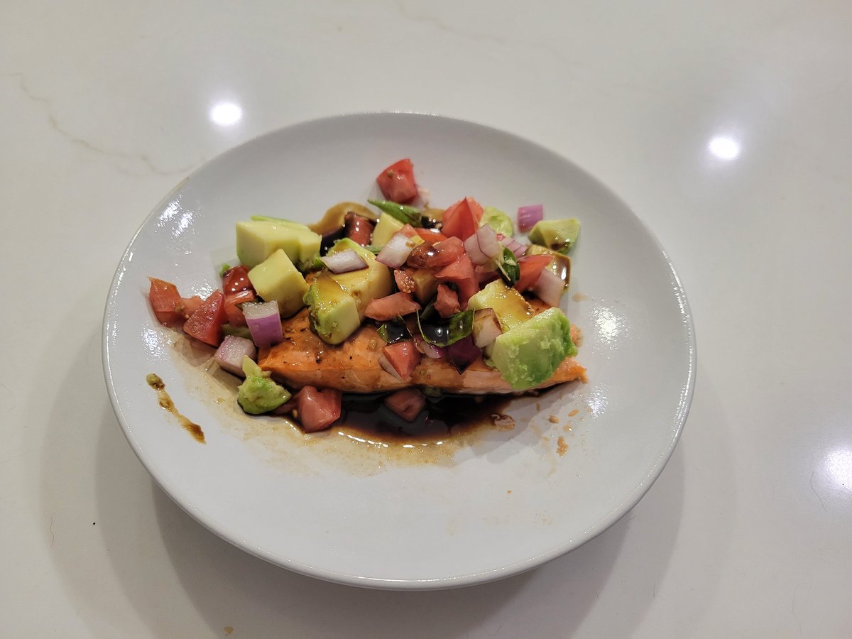 Baked salmon filet topped with diced tomato, red onion, basil and avocado, with balsamic vinegar pooled around the plate
