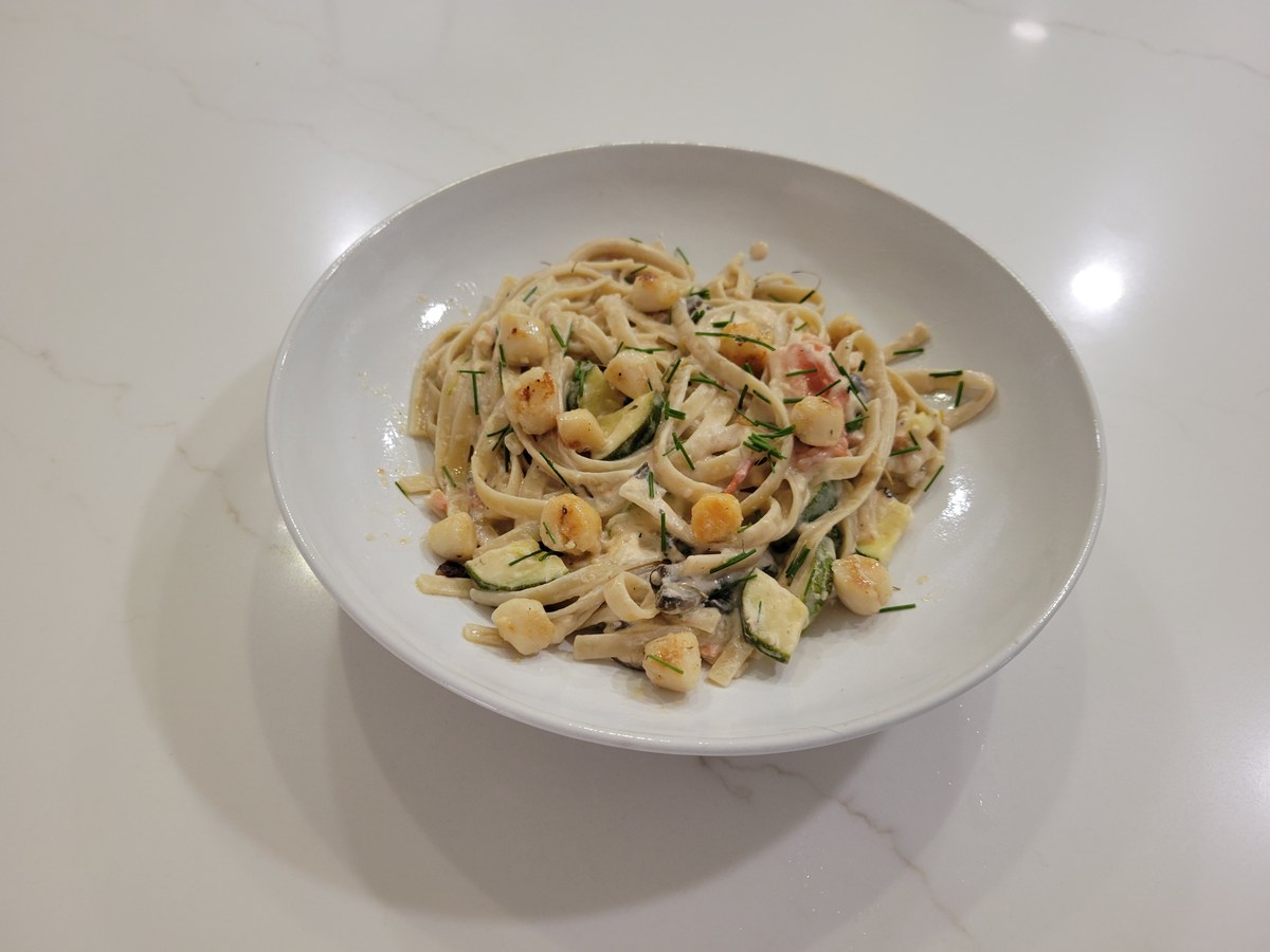 A shallow white bowl of linguine in a creamy lemon-Parmesan sauce, dotted with small seared bay scallops, zucchini chunks, mushroom, tomato, and a scatter of chives across the top.