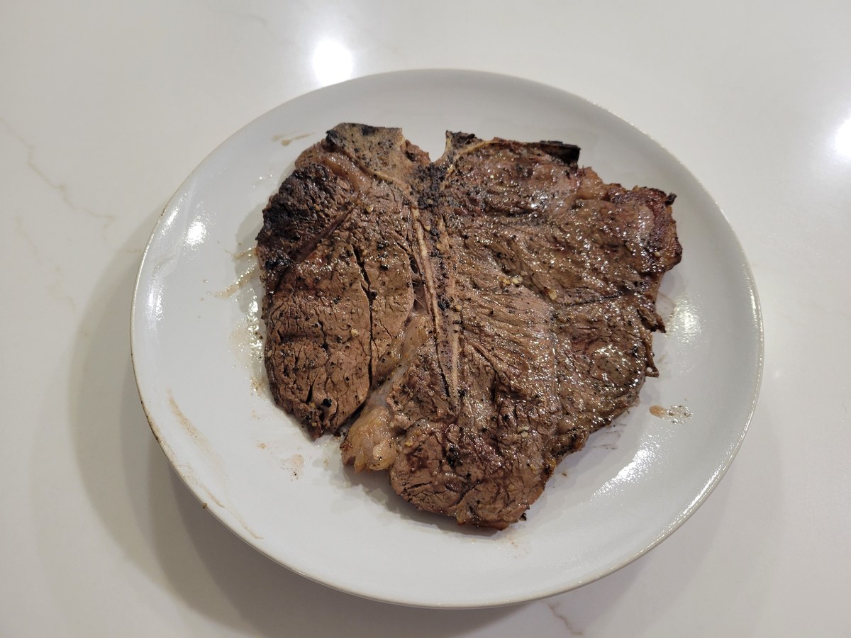 Whole porterhouse steak on a white plate, T-bone clearly visible with the strip loin on one side and tenderloin on the other, deep sear and char on the exposed side, glossy juice pooling on the plate after resting