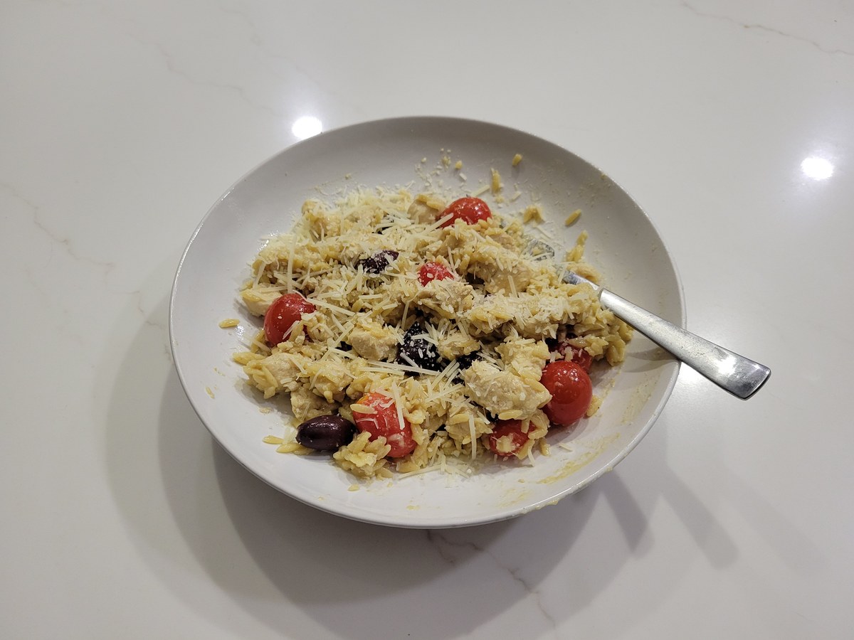 A bowl of chicken orzo with halved cherry tomatoes, kalamata olives, diced chicken, and grated Parmesan on top.