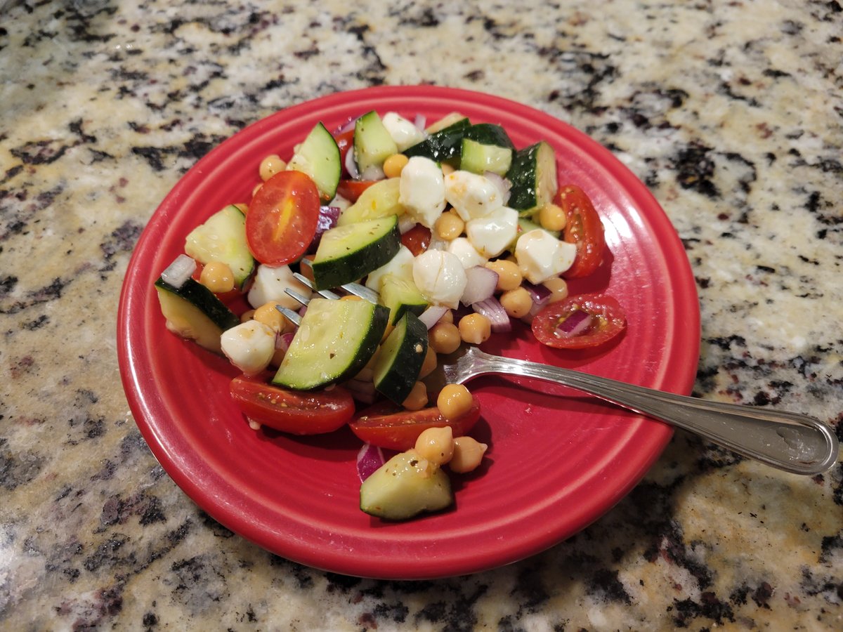 A red plate piled with chunky cucumber, halved grape tomatoes, fresh mozzarella pearls, chickpeas, and diced red onion, glossy with Italian dressing, fork stuck in the pile