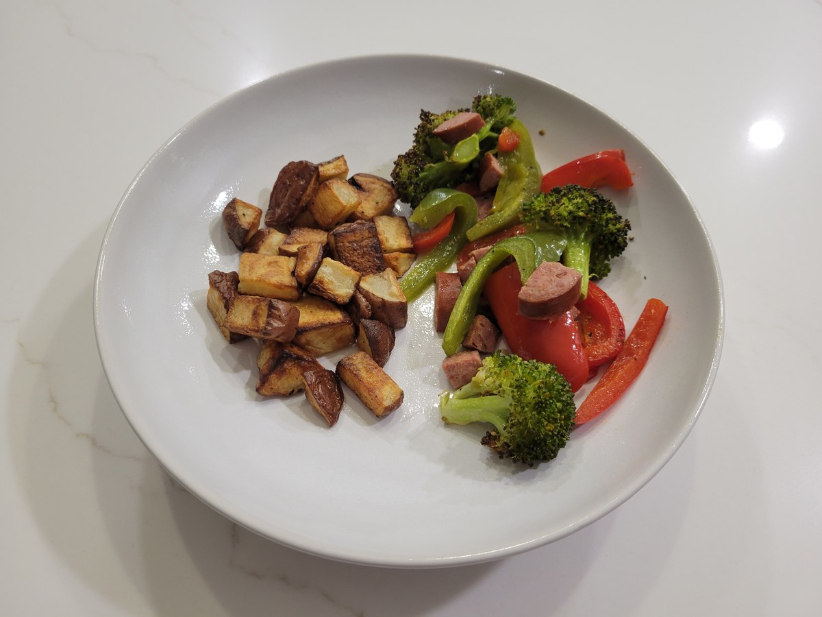 Wide shallow white bowl on marble: crispy baby red potato cubes piled on the left, sausage rounds with charred broccoli florets and red and green bell pepper strips on the right