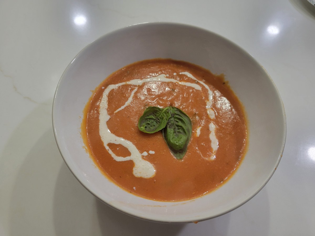 A wide shallow bowl of glossy orange-pink tomato bisque with an S-swoosh of heavy cream drizzled across the middle, three fresh basil leaves clustered in the center as garnish, sitting on a marble counter.